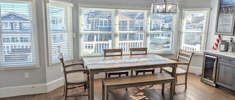 A dining area with a large wooden table and benches, surrounded by large windows with blinds, a chandelier, and a view of a building outside.