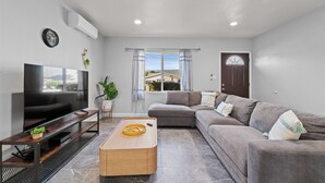 A modern living room with a flat-screen TV, a wooden coffee table, a grey sectional sofa, and a large window with curtains. - Tranquil 2-bedroom house with AC in pleasant Rosemead (Rosemead)