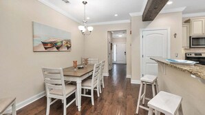 A dining area with a wooden table and white chairs, a painting on the wall, and a kitchen with a microwave and cabinets. - Close to Island Amenities (St. Simons Island)