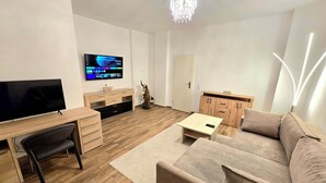 A modern living room with a flat-screen TV, wooden furniture, a beige sofa, and a chandelier. - Executive apartment + terrace + private parking space (Chemnitz)