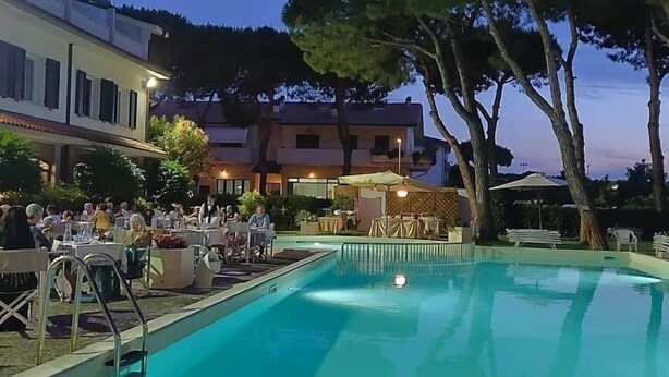 A swimming pool with a deck area, surrounded by trees and buildings. - Villa Elprado (Cervia)
