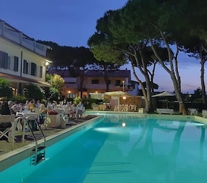 A swimming pool with a deck area, surrounded by trees and buildings. - Villa Elprado (Cervia)