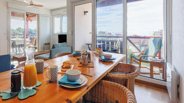 A dining area with a wooden table set for breakfast, wicker chairs, and a view of the city through large windows. - Appartement Paloma, vue océan - Welkeys (Soorts-Hossegor)