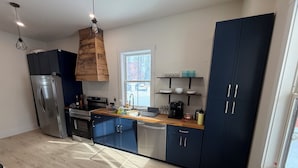 Una cucina moderna con frigorifero in acciaio inox, forno integrato, lavastoviglie e mobili in legno. Una finestra offre luce naturale e un lampadario illumina il piano di lavoro.