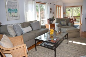 Ein Wohnzimmer mit einer Couch, Sesseln, einem Glastisch und einem Essbereich mit Tisch und Stühlen.