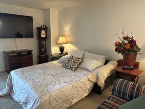 Un dormitorio con una cama grande, una mesita de noche de madera, una lámpara, un reloj, un televisor y un arreglo floral.