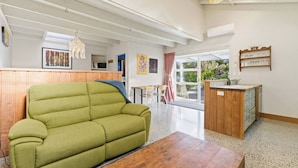 Ein geräumiges Zimmer mit einem grünen Sofa, einem Holztisch, einer kleinen Küchenzeile mit Spüle und Regalen und einem großen Fenster mit Blick ins Freie.
