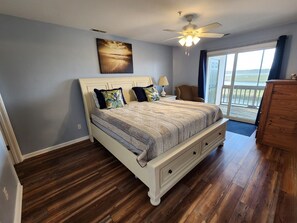 A bedroom with a bed, a ceiling fan, a view of the outdoors, and a wooden floor.