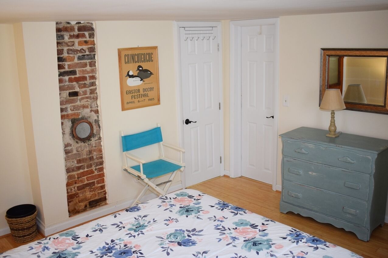 A bedroom with a brick wall, a blue folding chair, a white door, a dresser, and a bed with a floral bedspread.