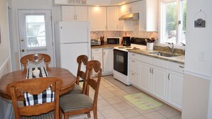 Une cuisine compacte avec des meubles blancs, un réfrigérateur blanc, un four à micro-ondes, une cuisinière et un évier. On y trouve une table à manger avec des chaises et une fenêtre donnant sur l’extérieur.