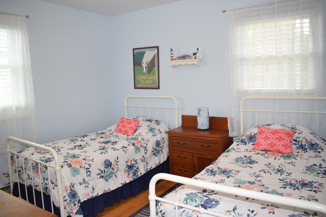 Un dormitorio con dos camas individuales, un aparador de madera, un cuadro enmarcado en la pared y una ventana con cortinas de encaje.
