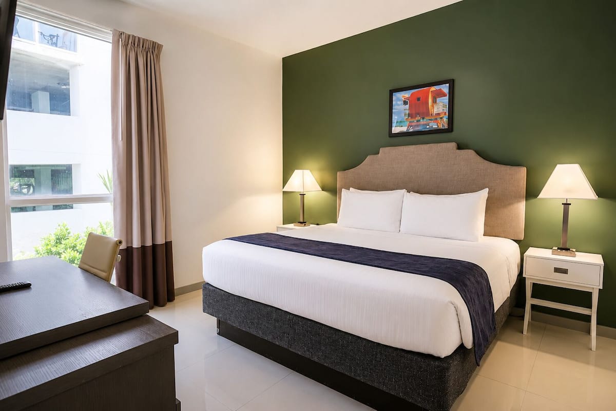 A hotel room with a bed, two bedside tables with lamps, a desk with a remote control, and a window with curtains.