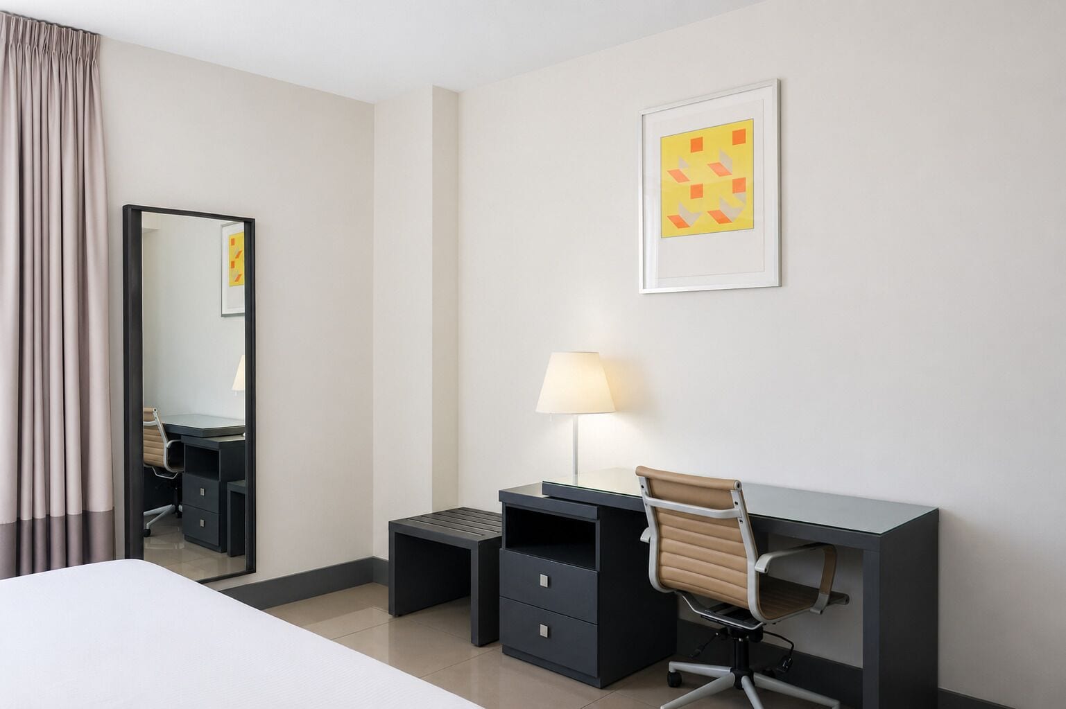 A hotel room with a bed, a desk with a chair, a lamp, a mirror, and a framed picture on the wall.