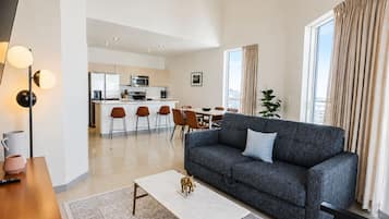 Una sala moderna con un sofá gris, una mesa de centro y un comedor con sillas, y una cocina al fondo.