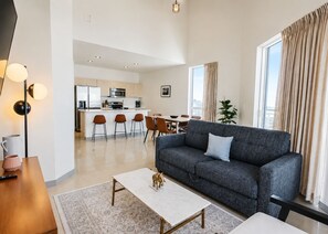 A modern living room with a grey sofa, a coffee table, and a dining area with chairs and a kitchen in the background. - Habitat Brickell (Miami)