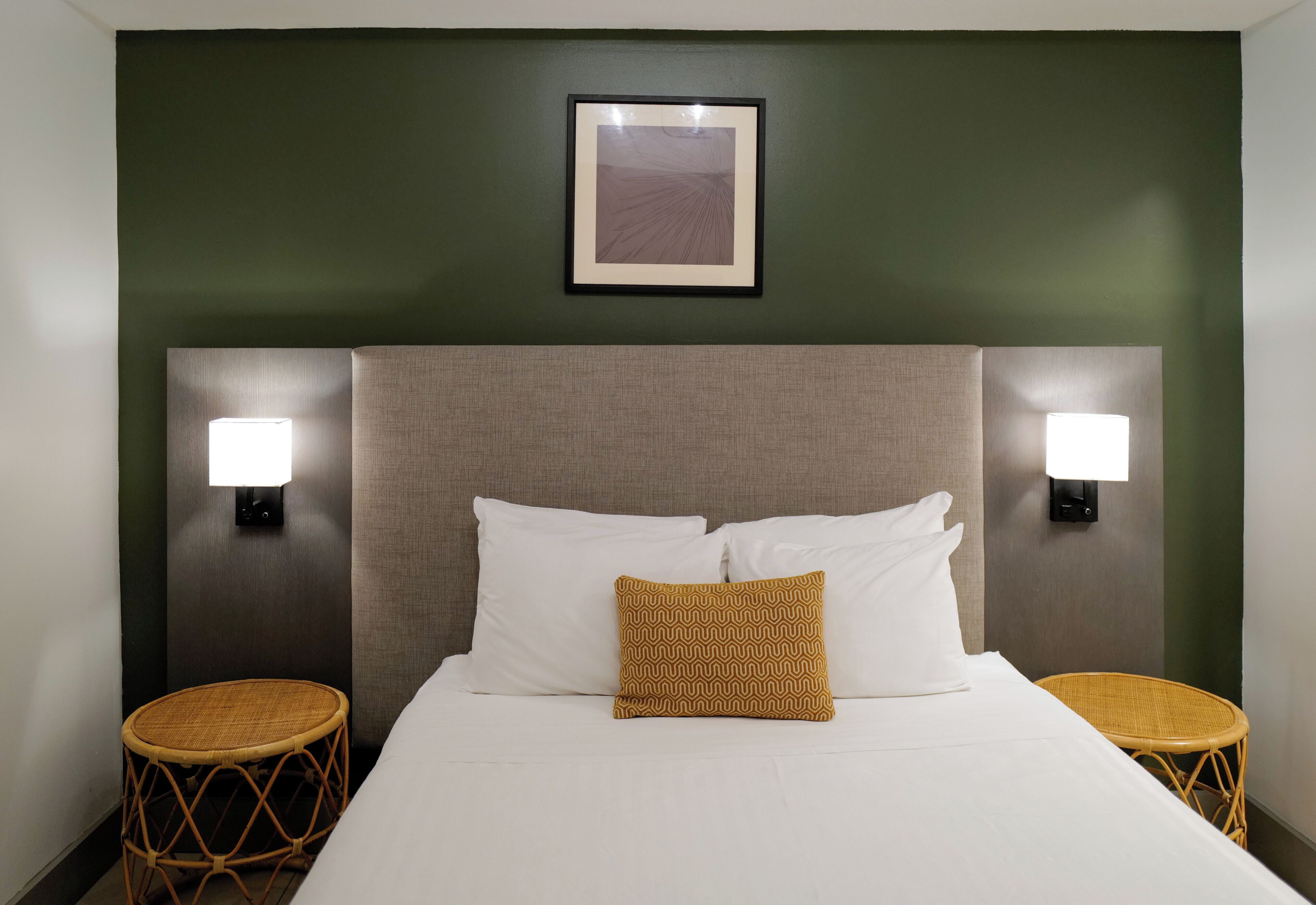 Een hotelkamer met een bed, twee nachtkastjes, twee wandlampen, een ingelijst schilderij boven het bed en twee rieten bijzettafeltjes.