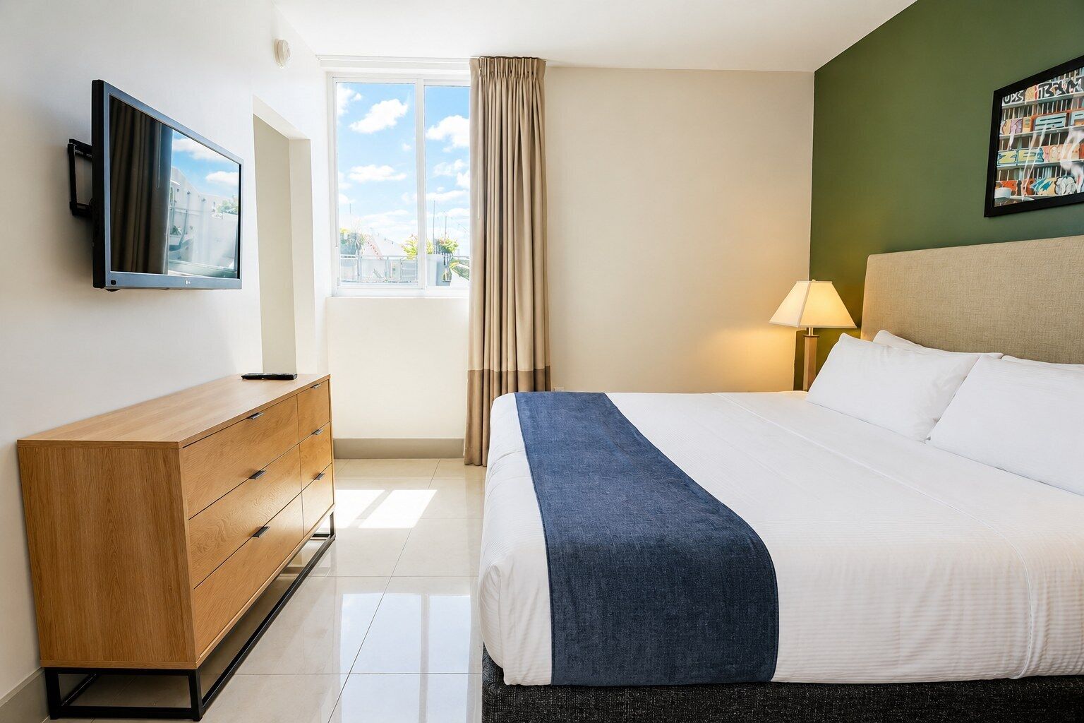 Een hotelkamer met een bed, een tv die aan de muur hangt, een dressoir, een raam met gordijnen en een lamp.