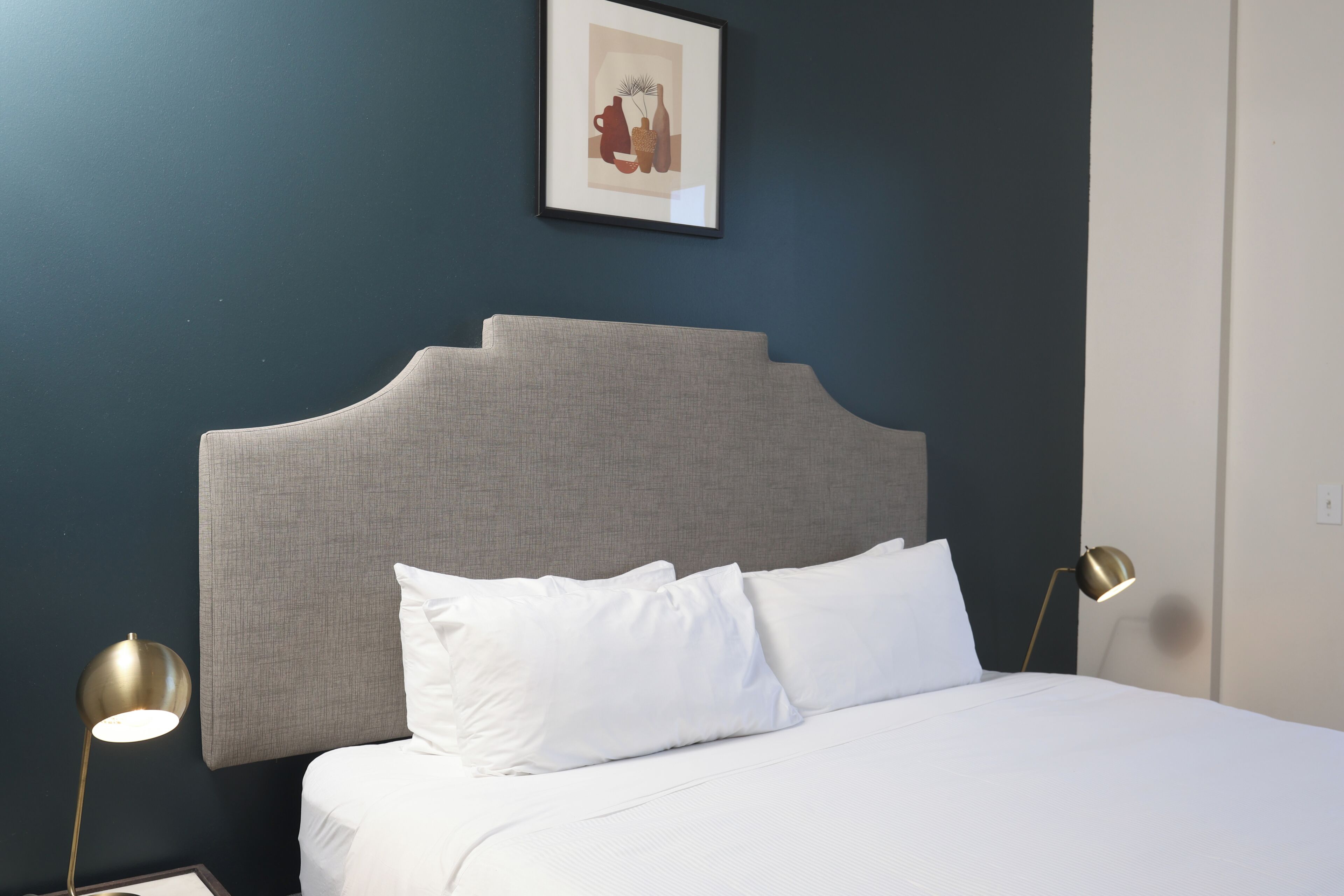 A neatly made bed with white linens, a grey upholstered headboard, and two bedside lamps.