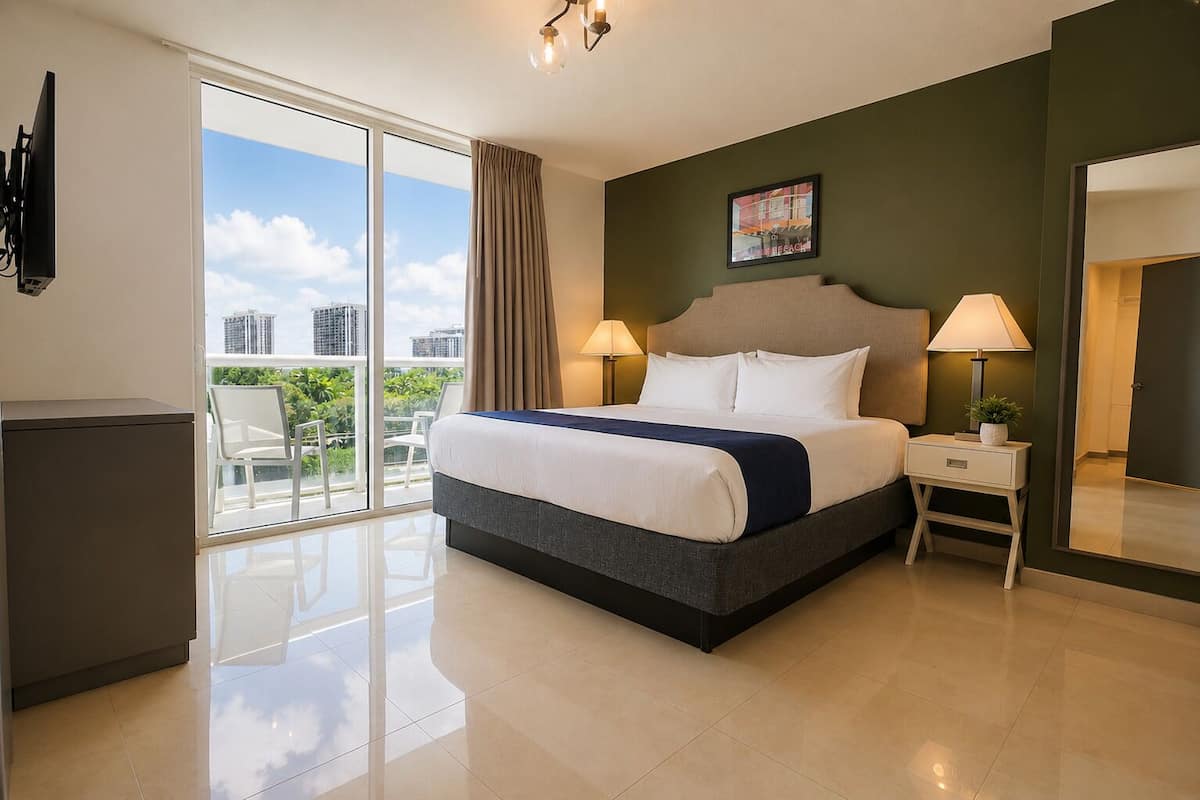 A hotel room with a bed, two bedside lamps, a nightstand, a mirror, and a view of the city through large windows.