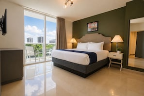 A hotel room with a bed, two bedside lamps, a nightstand, a mirror, and a view of the city through large windows.