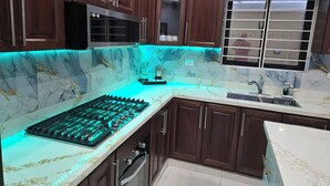 Una cocina con armarios de madera oscura, un microondas de acero inoxidable, una estufa a gas con iluminación LED y una ventana con persianas.