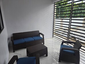 Un balcón con un sofá y una silla de mimbre, una mesita y vistas a zonas verdes a través de barrotes de metal.
