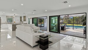 A modern living room with a white sofa, a glass coffee table, and a view of a swimming pool through large sliding glass doors. - 6 bedroom Oasis with Extra Large Pool, spa, EV Charger & Guest House, Sleeps 16+ (Naples)
