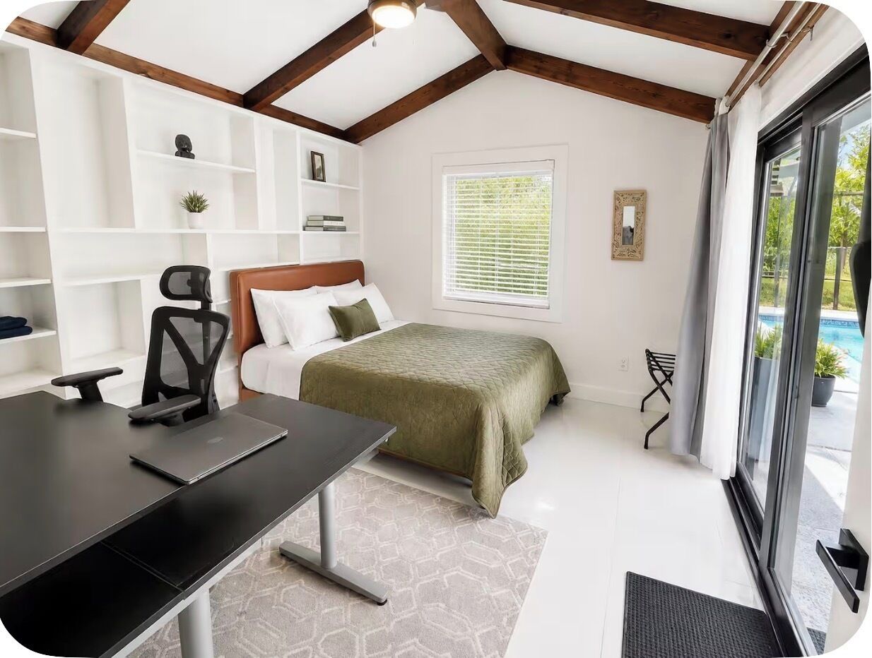 Une chambre avec un lit, un bureau et une chaise. Il y a une fenêtre avec des stores, une porte et une vue sur l’extérieur.