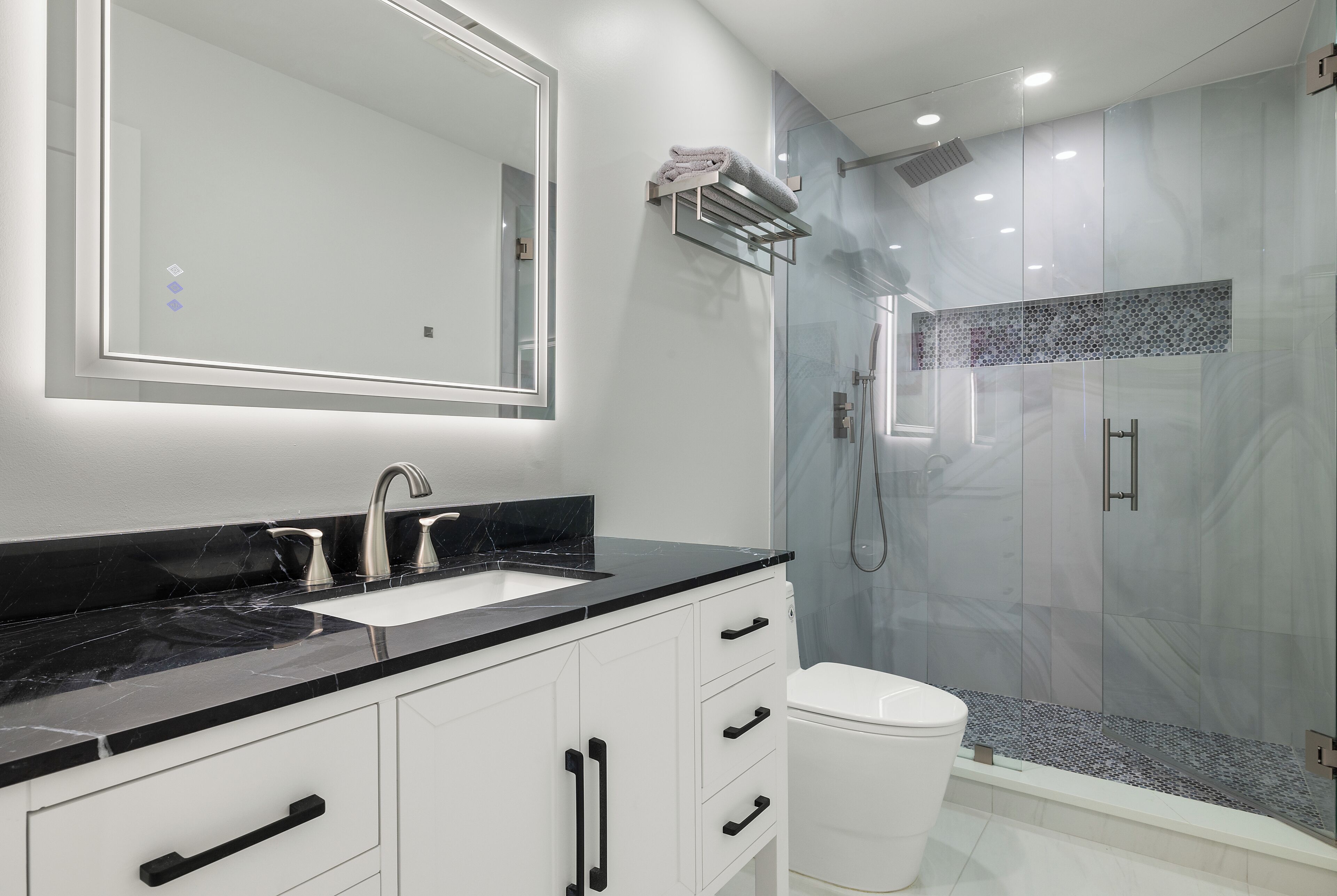 Un bany modern amb un mirall gran, una superfície de lavabo negra, un lavabo blanc i una dutxa amb envàs de vidre.