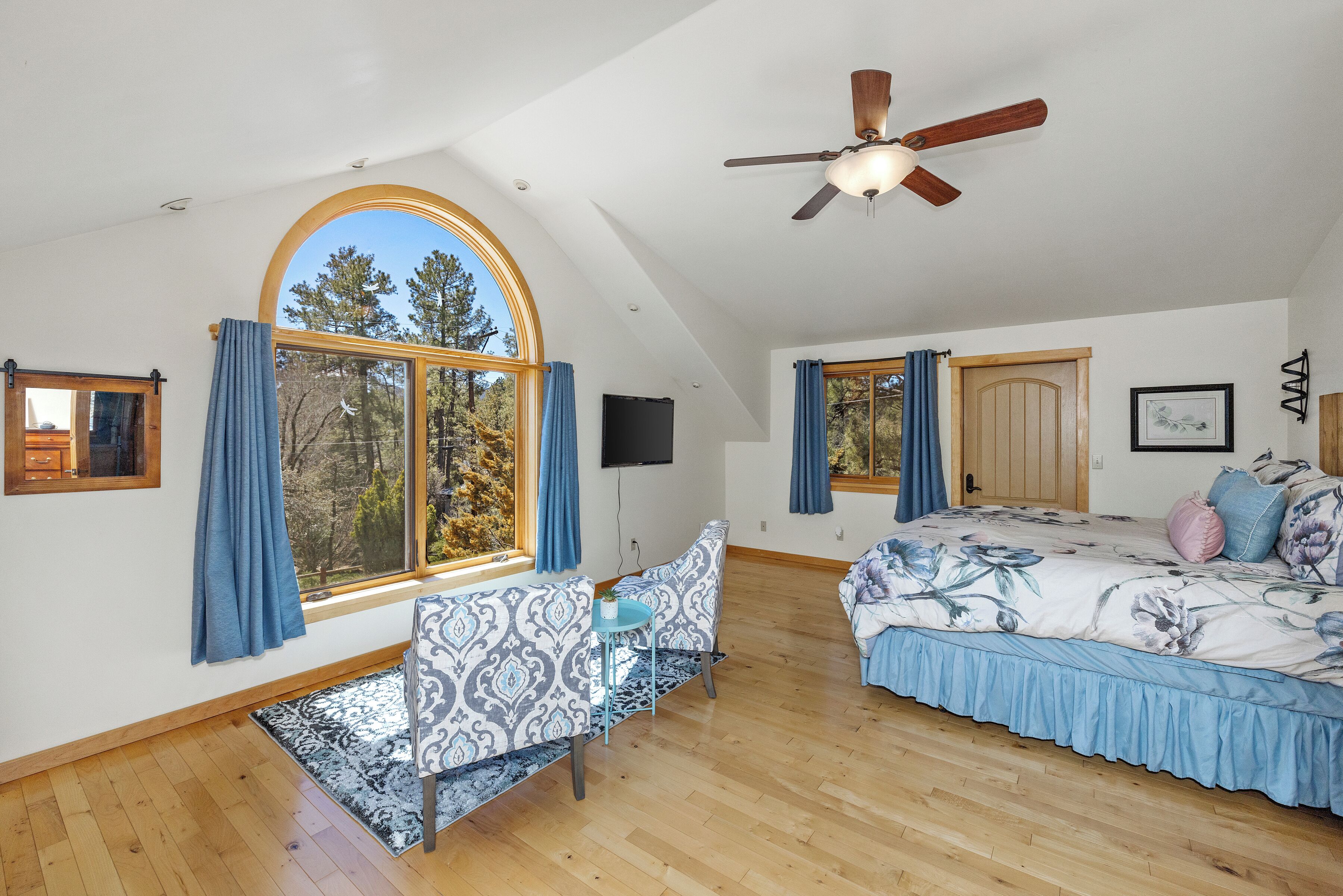 A spacious room with a large arched window, a ceiling fan, a bed with floral bedding, a sofa with patterned upholstery, and a small table with a plant.