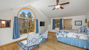 A spacious room with a large arched window, a ceiling fan, a bed with floral bedding, a sofa with patterned upholstery, and a small table with a plant.