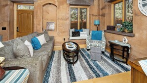 Una sala de estar acogedora con una puerta de madera, un sofá, un sillón estampado, una mesita de centro y una ventana con vistas a árboles.