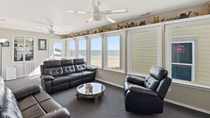Un salon avec un canapé en cuir noir, une table basse en verre et un fauteuil noir. Il y a des ventilateurs de plafond et, grâce à de grandes fenêtres, on peut admirer la vue sur la plage.