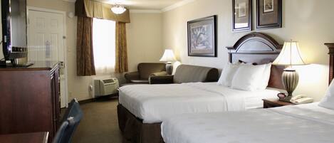 A hotel room with two beds, a desk, a chair, a TV, and a lamp.