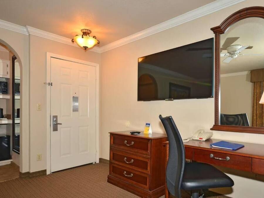 Quarto de hotel com porta branca, cómoda de madeira, escrivaninha com cadeira, espelho grande e ventoinha no teto.