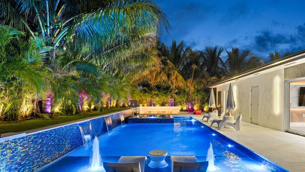 Una piscina con cascada, rodeada de palmeras y mobiliario de exterior.
