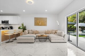 Ein modernes Wohnzimmer mit einem Ecksofa, einem Couchtisch und einer Küchenzeile mit weißen Schränken und einer Mikrowelle.