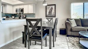 Una cocina moderna con armarios blancos, una zona de comedor con mesa y sillas de madera oscura, y una sala de estar con sofá beige y alfombra estampada.