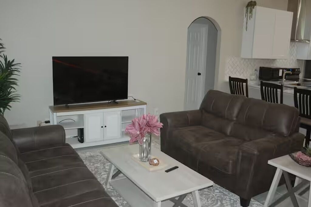Un salon avec un canapé en cuir marron, une table basse blanche ornée d’un vase rose et d’une télécommande, et un téléviseur à écran plat posé sur un meuble blanc.
