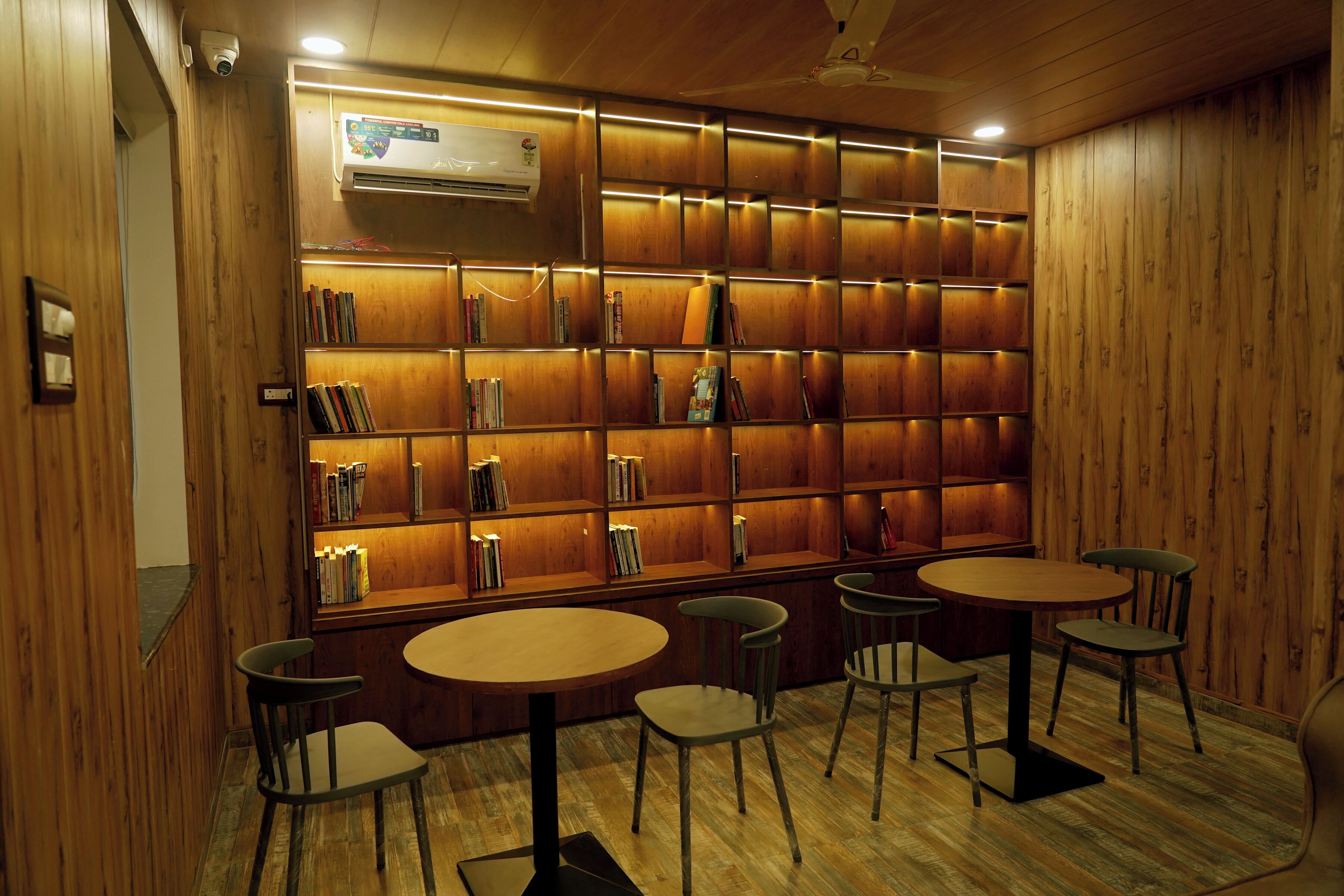A library with wooden bookshelves, a ceiling fan, and four tables with chairs.