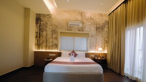 A hotel room with a large bed, a window with blinds, an air conditioning unit, a bedside table with a lamp, and a wall with a tree mural. - The One Jubilee (Hyderabad)