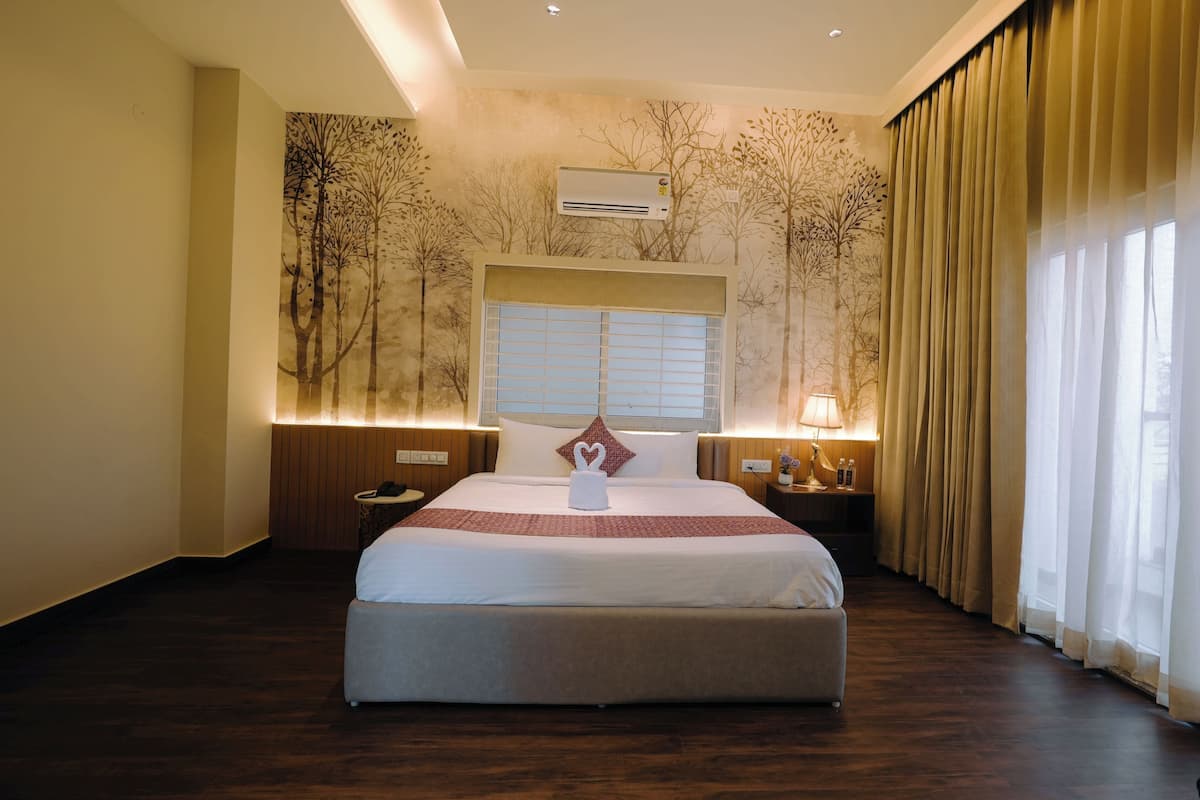 A hotel room with a large bed, a window with blinds, an air conditioning unit, a bedside table with a lamp, and a wall with a tree mural.