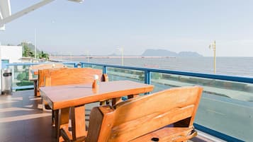 Restaurante à beira-mar com mesas e cadeiras de madeira, guarda-corpo de vidro e vista para o mar e montanhas ao longe.