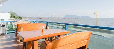 A seaside restaurant with wooden tables and chairs, a glass railing, and a view of the sea and distant mountains.