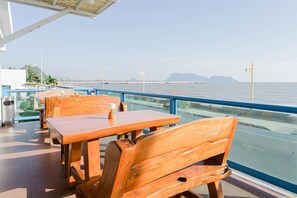 A seaside restaurant with wooden tables and chairs, a glass railing, and a view of the sea and distant mountains. - Hadthong Hotel (Prachuap Khiri Khan)