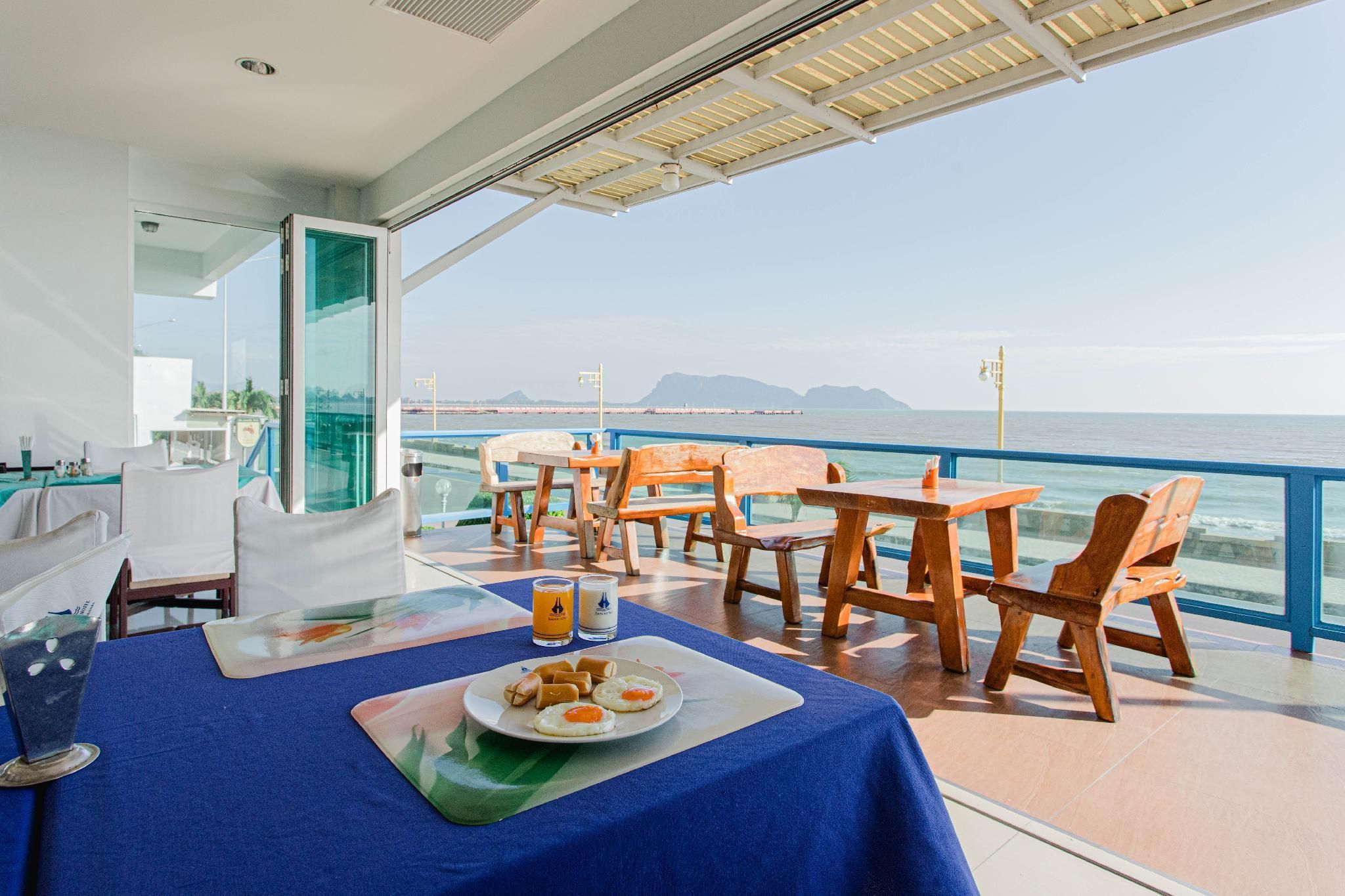 A dining area with a view of the sea, featuring a table with a plate of food and drinks, wooden chairs, and a glass partition.