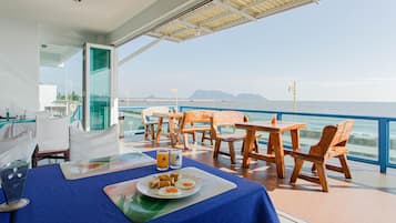 Uma área de refeições com vista para o mar, com uma mesa, prato de comida e bebidas, cadeiras de madeira e uma divisória de vidro.