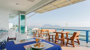 A dining area with a view of the sea, featuring a table with a plate of food and drinks, wooden chairs, and a glass partition. - Hadthong Hotel (Prachuap Khiri Khan)