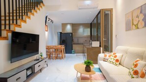 Una sala de estar moderna con un televisor de pantalla plana, un sofá con cojines estampados color naranja, una zona de comedor con sillas de madera, una escalera y una cocina con refrigerador y armarios.