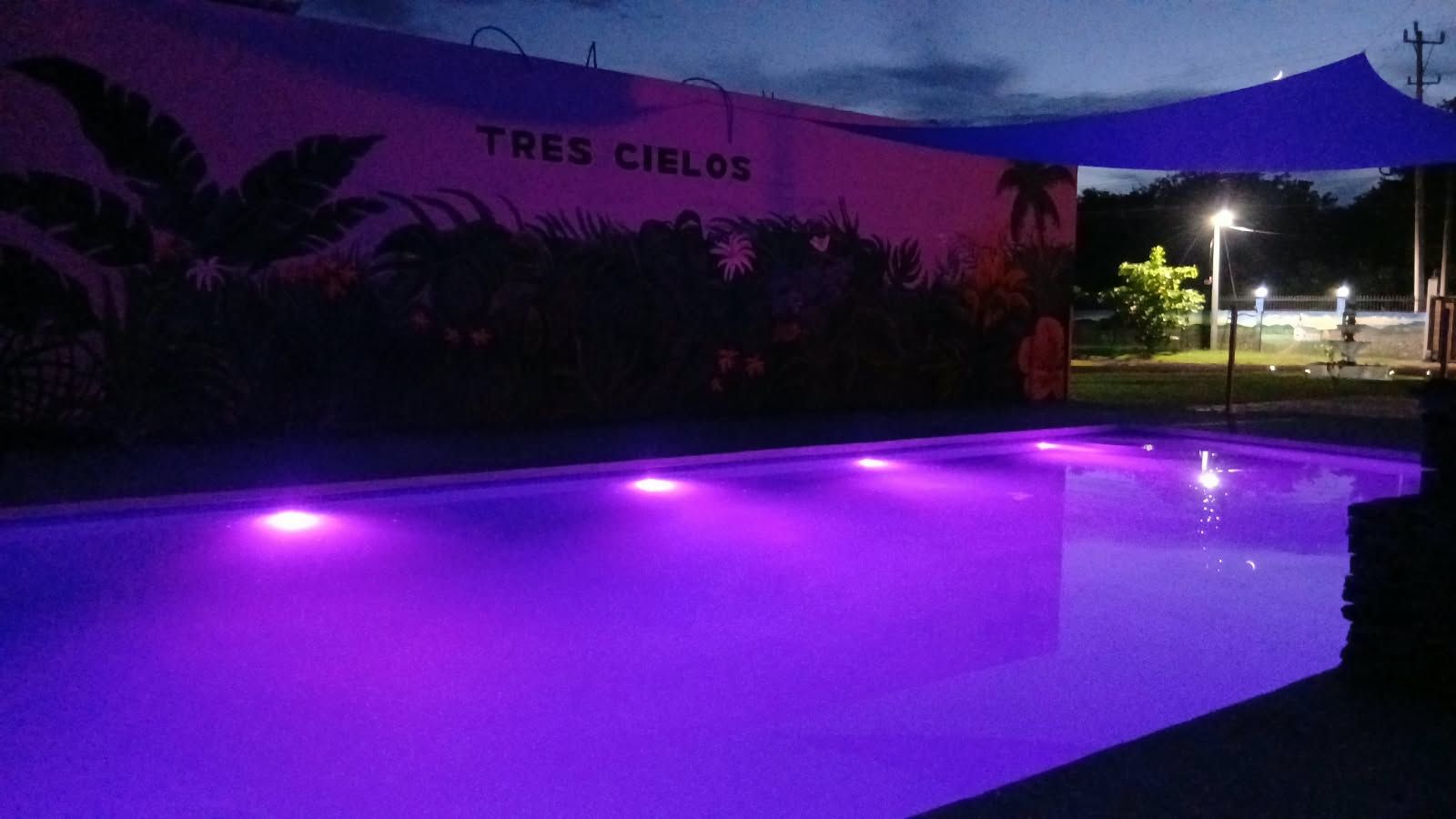 A swimming pool with purple lighting, surrounded by a wall with tropical plants and the text 'TRES CIELOS'.